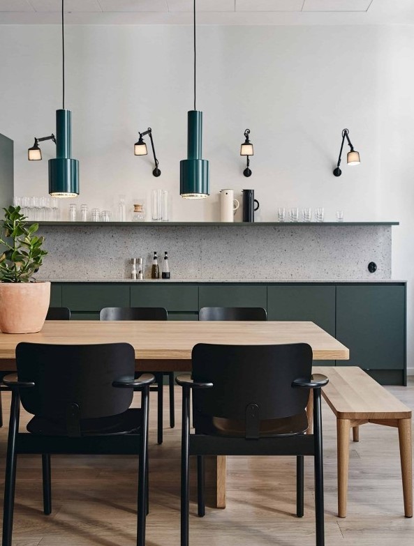 a modern kitchen with sleek no hardware cabinets, a terrazzo backsplash, a long open shelf, green pendant lamps and cool sconces, a blonde wood table and a bench, black chairs