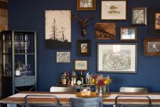 a moody dining room with a navy accent wall with a gallery wall in woodland style, a planked table and black chairs