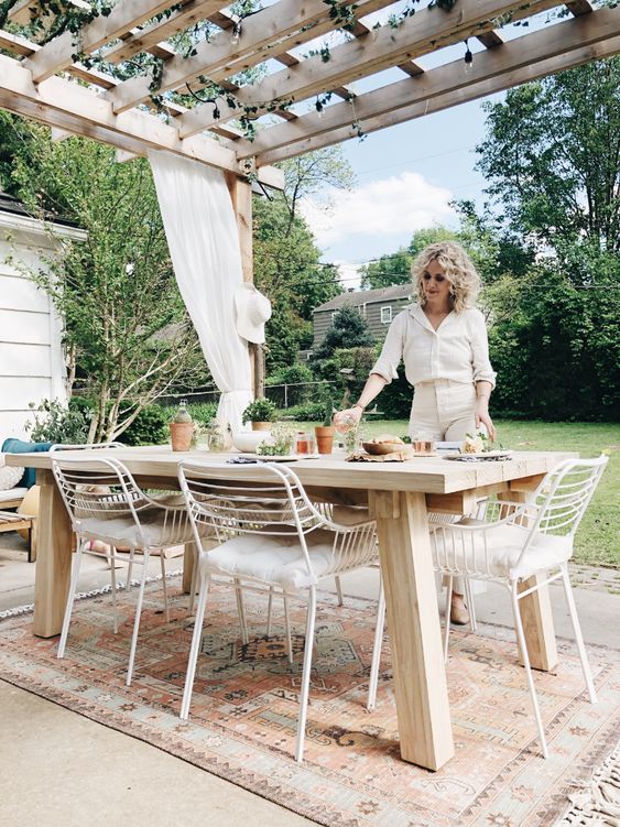 a neutral boho space with a roof with greenery and neutral curtains, a light-stained table and white metal chairs plus a garden view