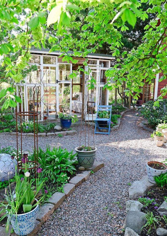 a pretty and cute garden with greenery and blooms in garden beds with brick and rock edges, gravel paths, a former greenhouse turned into an outdoor room