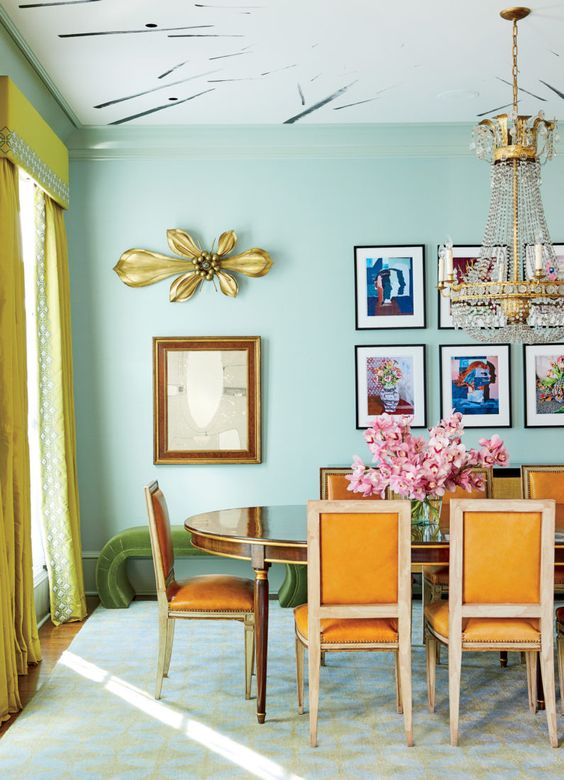 a quirky colorful dining room with mint green walls, a chic polished table and orange chairs, a statement chandelier and a grid colorful gallery wall