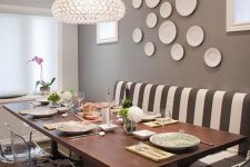 a quirky taupe dining room with a striped sofa, a wooden table, acrylic chairs, a chic chandelier and a gallery wall of plates