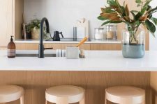 a sleek minimalist blonde wood kitchen with white countertops and a backsplash, black fixtures and some magnolia leaves