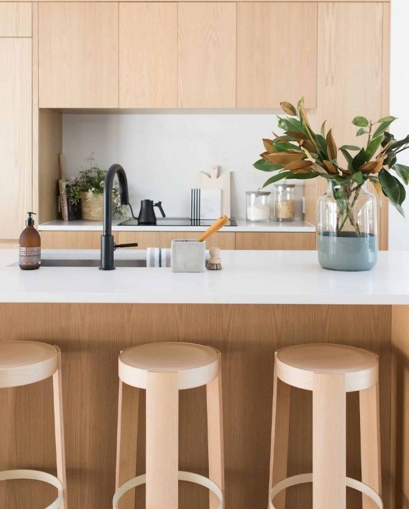 a sleek minimalist blonde wood kitchen with white countertops and a backsplash, black fixtures and some magnolia leaves