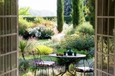 a small yet chic Provence outdoor space with a metal round table and forged chairs with striped cushions and lots of greenery and blooms around
