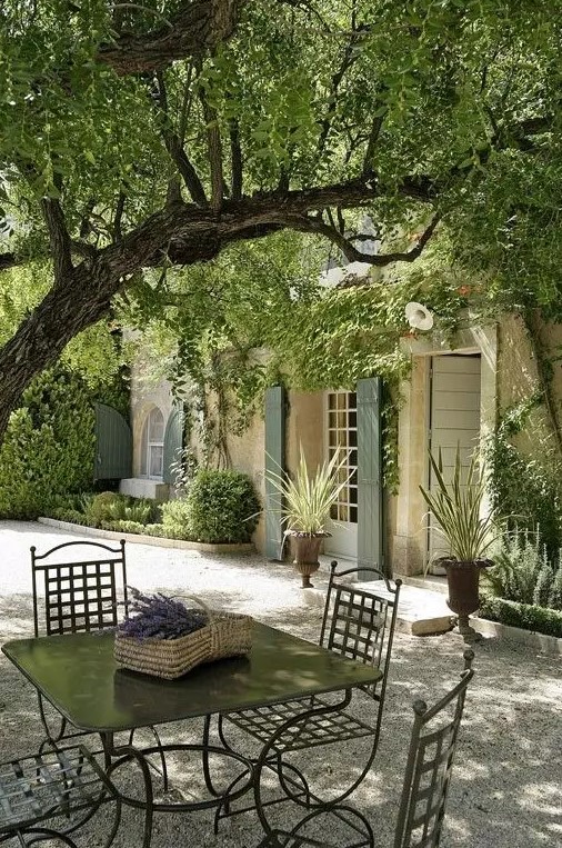 a small yet very simple and chic Provence terrace with dark metal furniture, a tree over the space and potted greenery