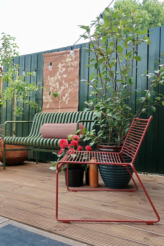 a stylish and bright terrace with a tall fence in green, a green metal sofa and a red chair, some potted plants and greenery