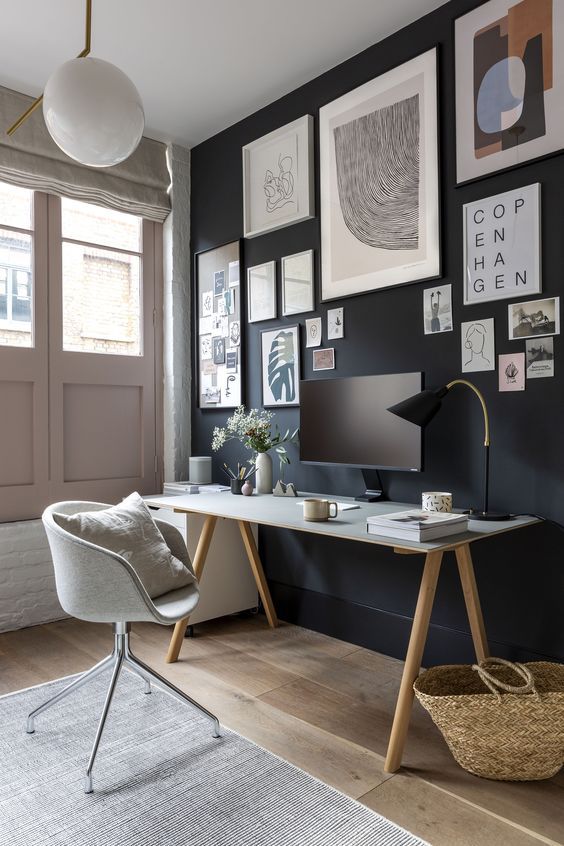 a stylish home office with a black accent wall with a gallery wall, a desk, a grey chair, a white cabinet and a pendant lamp