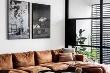 a stylish living room with a rust sectional, a grey chair, black round tables, a black and white gallery wall and some potted plants