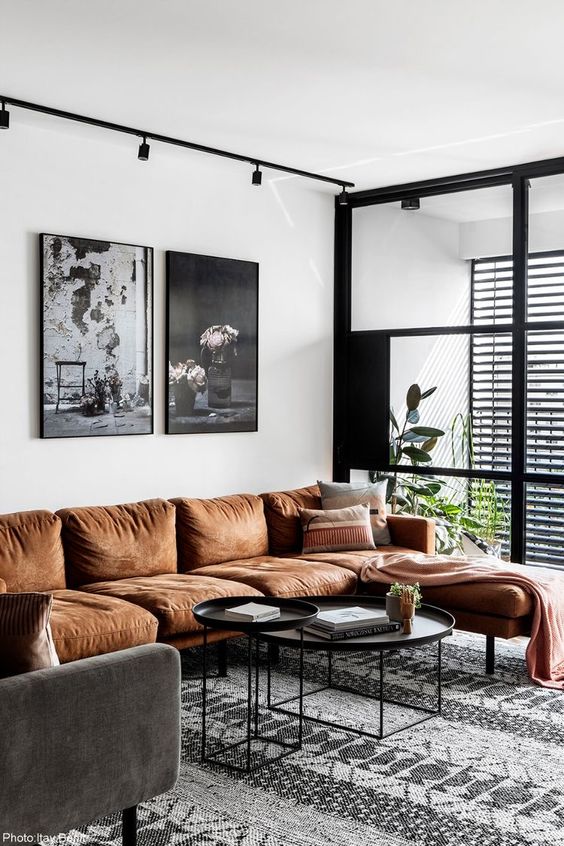 a stylish living room with a rust sectional, a grey chair, black round tables, a black and white gallery wall and some potted plants