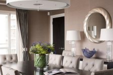 a taupe dining room with a black table and grey leather chairs, a large pendant lamp, curtains and a round mirror