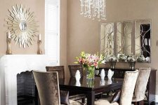 a taupe living room with a non-working fireplac,e a black table, taupe chairs, a chic crystal chandelier and a mirror of three parts