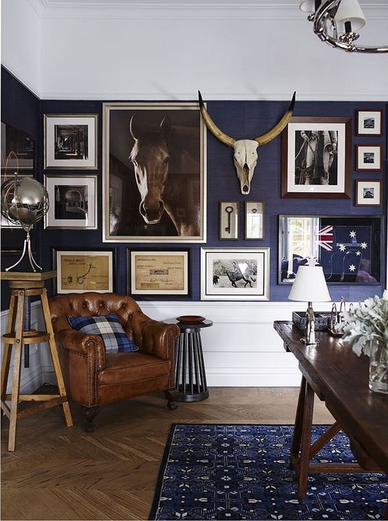a vintage rustic home office with a stained desk, a leather chair, a large gallery wall on a cobalt wall and white paneling