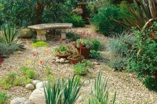 a wild-looking garden with lot sof greenery and shrubs, with potted plants and a stone bench, with gravel pathways