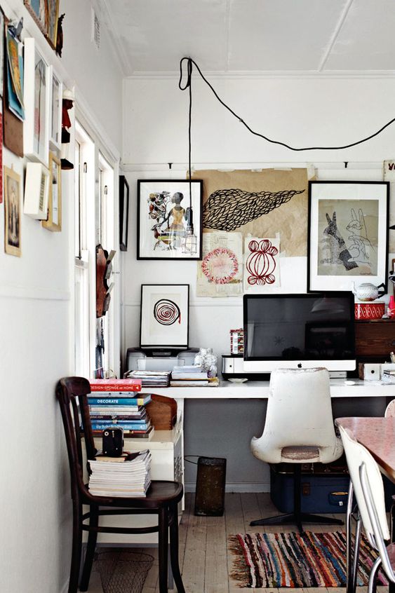 an eclectic home office wiht a white desk and a creamy chair, with amessy gallery wall that extends to the next wall, some chairs and a bold rug