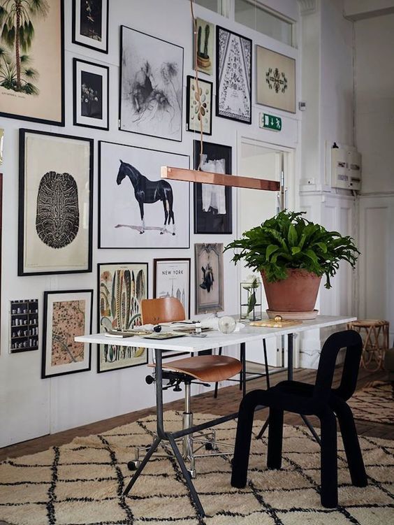 an eclectic home office with a lightweight desk, a plywood and black soft chair, an oversized gallery wall with potted plants