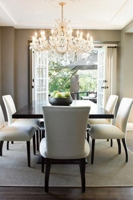 an elegant taupe dining room with a large dark stained table, creamy chairs, a neutral rug and an oversized chandelier plus a garden view