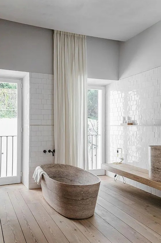 a minimalist neutral bathroom with grey walls, white subway tiles, a wooden floating bench and a stone tub plus windows and curtains under the ceiling to highlight the double height