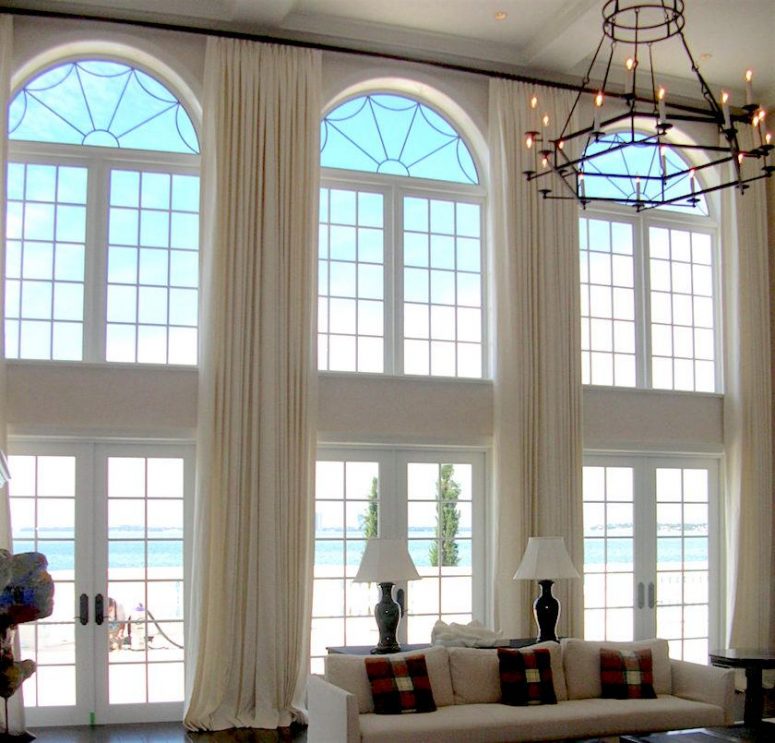 a modern living room with tan furniture, double-height ceiling and windows with arched tops done with matching neutral drapes that cover everything
