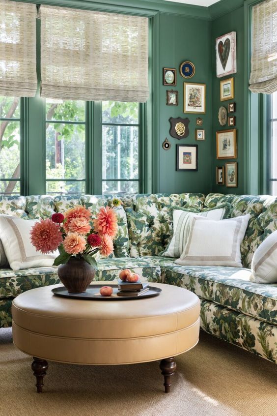 a 1920s inspired living room with green walls, a botanical print corner sofa, a beige round pouf, a corner gallery wall and bright blooms