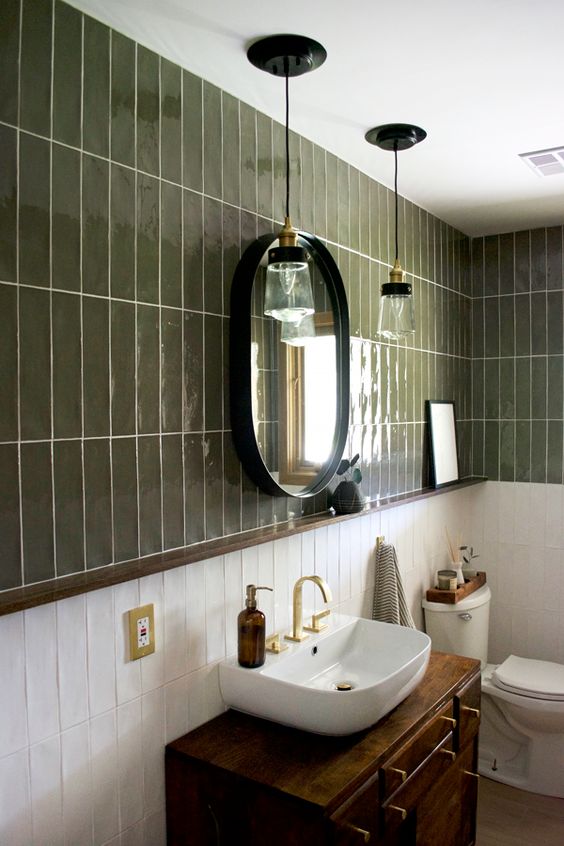 a beautiful and elegant bathroom with green and white stacked tiles, a dark stained vanity with a sink, an oval mirror and pendant lamps
