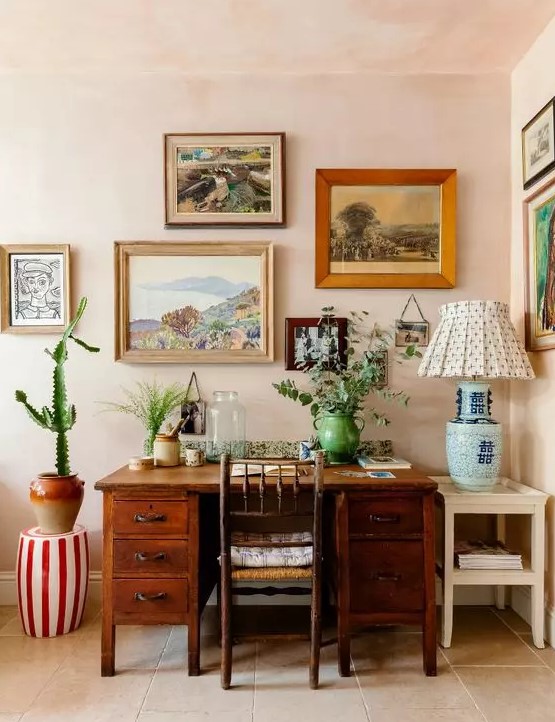a bright mid-century modern home office with blush walls, a stained desk and a chair, a colorful gallery wall and greenery and a cactus