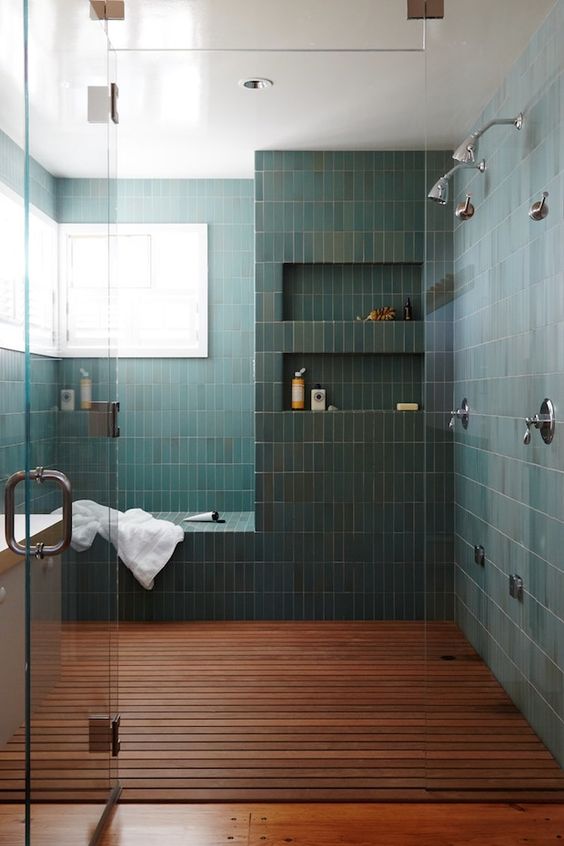 a chic bathroom with a large shower space clad with green stacked tiles, with a bench and niches in the walls, a dark stained floor and windows for natural light