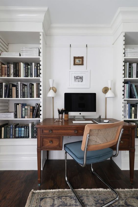 a chic home office with white built-in bookcases, a stained vintage desk, a modern chair and some art and chic lamps is a cool space