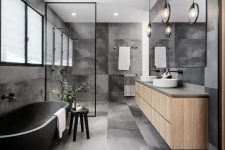a contemporary grey bathroom clad with concrete tiles, a stone bathtub, a floating vanity, round sinks, black fixtures and framing