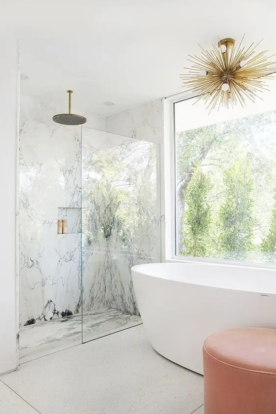 a fancy bathroom with white marble in the shower space, a glazed wall, a bathtub, a coral ottoman and a gold sunburst chandelier