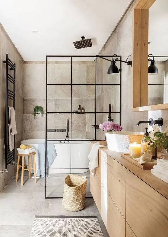 a greige bathroom clad with concrete, with a light stained vanity and a large mirror, a shower space with a black space divider and black fixtures
