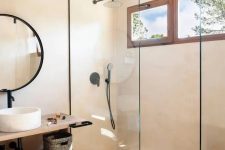 a greige contemporary bathroom with a shower space enclosed in glass, a floating open shelf vanity, a round mirror in a black frame, black fixtures