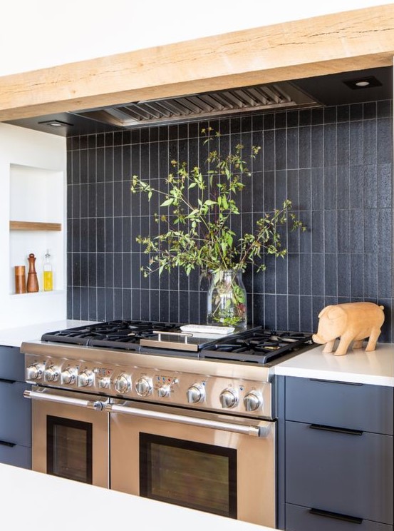 a grey kitchen with wooden beams, a black matte stacked tile backsplash and a metal cooker and hood