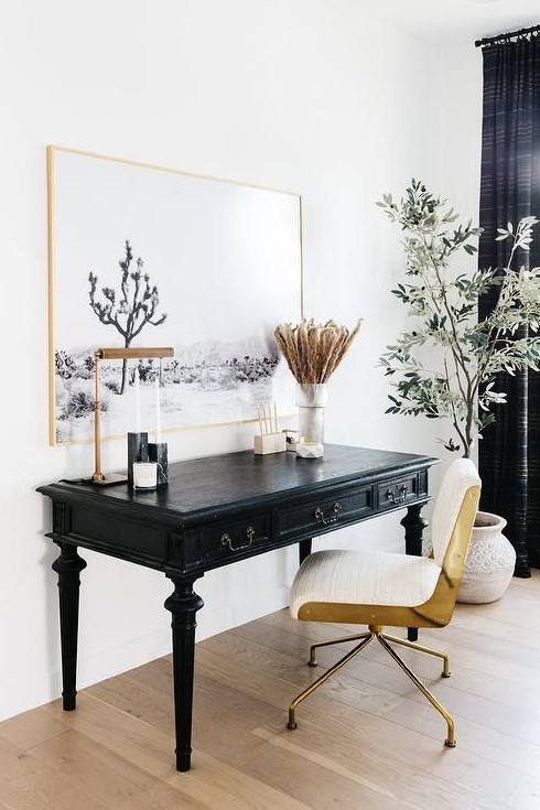 a lovely home office space with a black vintage desk, a white and gold chair, a statement artwork in a gold frame and black curtains