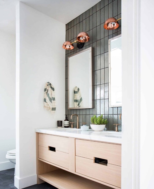 a modern bathroom with grey stacked skinny tiles, a blonde wood vanity with a white countertop, brass fixtures and copper sconces