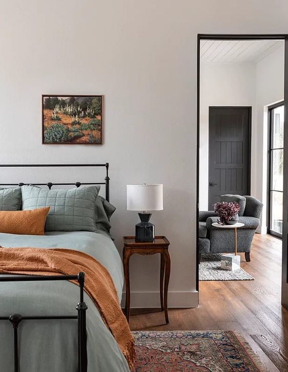 a modern bedroom with a black forged bed with muted green bedding, a dark-stained vintage nightstand and a table lamp