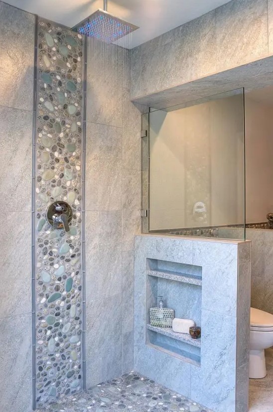 a neutral bathroom clad with tiles and faux pebbles, with a pony wall that had some shelves integrated is a lovely space