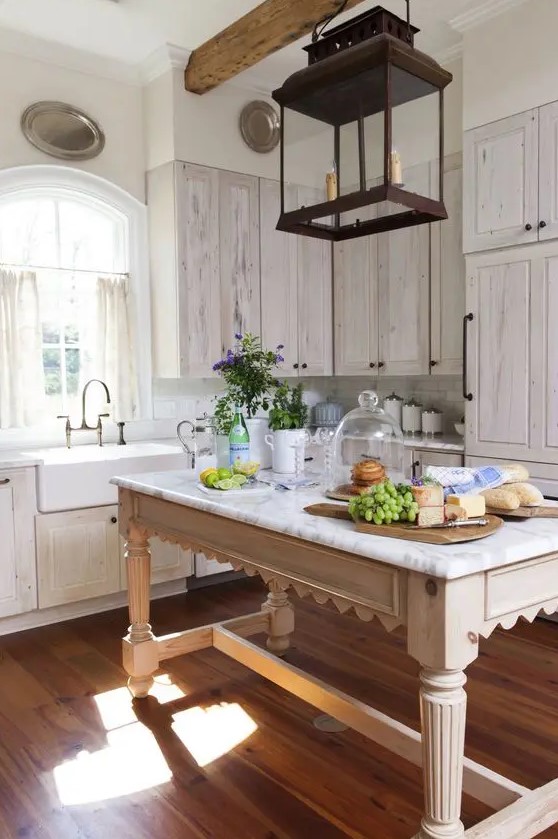 a pretty cottage whitewashed kitchen with white stone countertops, a neutral table that doubles as a kitchen island and a metal pendant lamp