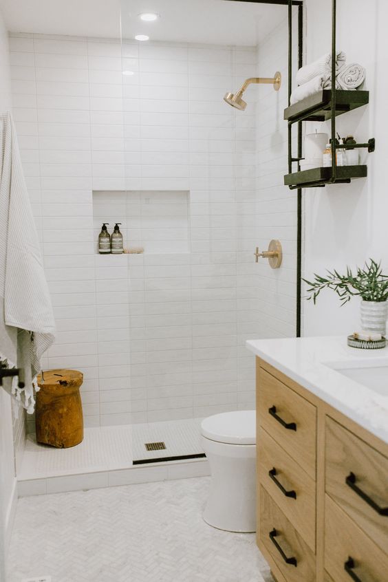 a small and cute neutral bathroom with white stacked tiles, herringbone marble ones, a blonde wood vanity, open shelving and a cork stool