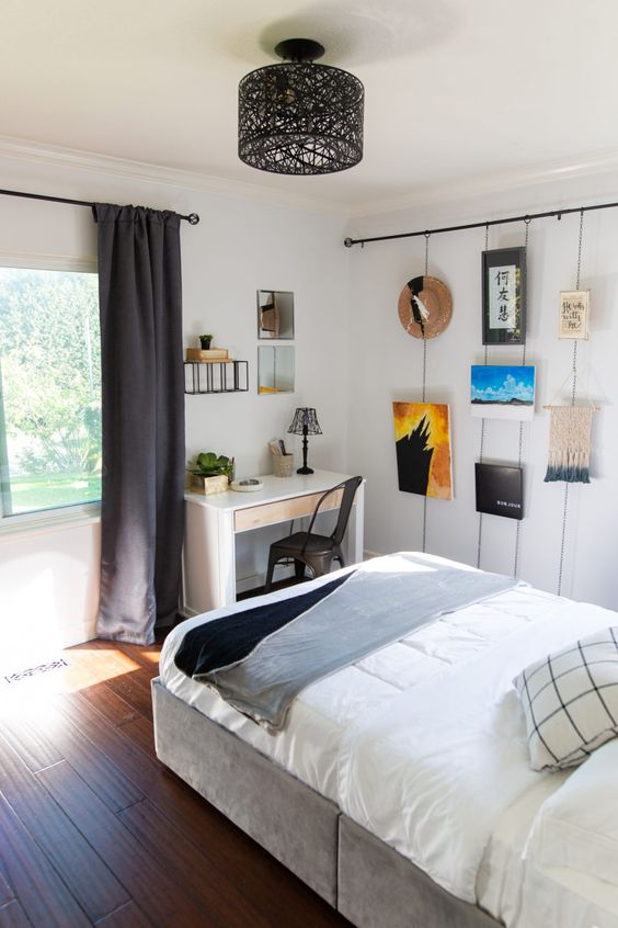 a small teen bedroom with a grey bed with neutral bedding, a white desk and a black chair, a gallery wall and dark curtains
