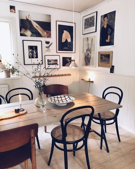 a stylish dining room with a bold corner gallery wall with mismatching frames, a stained table and black and neutral chairs, some lamps and plants