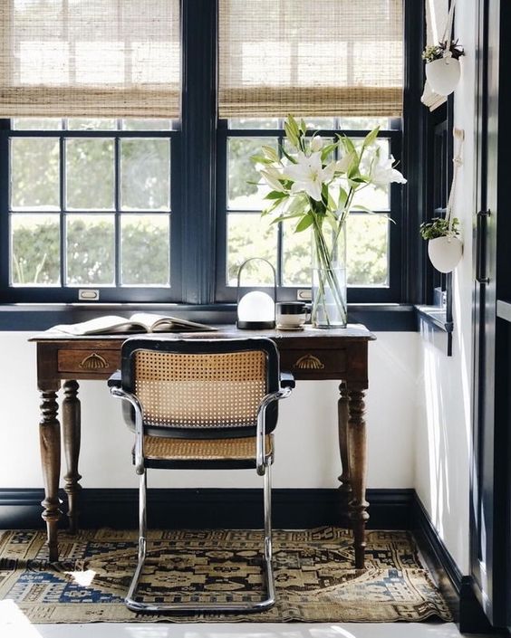 a stylish home office with black window frames, a vintage stained desk, a chic chair and a printed rug, neutral curtains