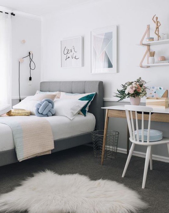 a stylish modern teen bedroom with a grey upholstered bed, pastel and color block bedding, a desk and a chair, some shelves and cool art