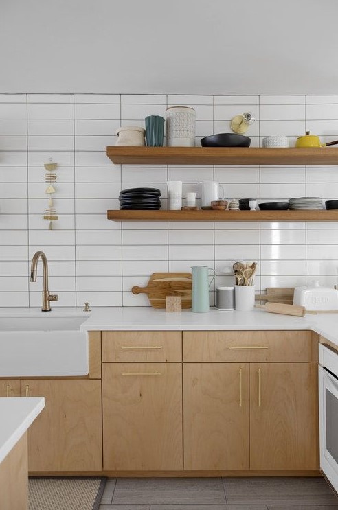 an airy Scandinavian kitchen with light colored wooden cabinets and shelves, a white stacked tile wall and brass fixtures
