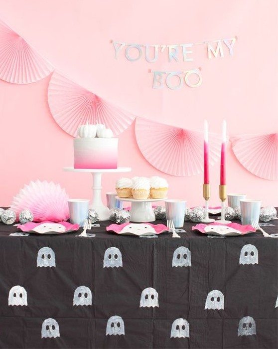 a pretty Halloween sweets table in pink and black, with pink paper fans, pink candles and a cake, pink ghost plates and a ghost tablecloth