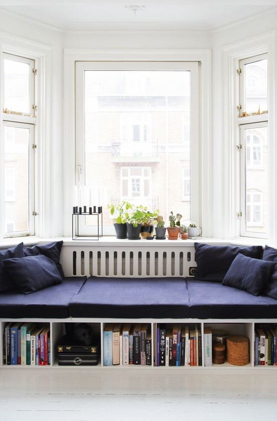 a bay window reading nook styled with midnight blue cushions and pillows, built-in bookshelves, potted plants and a candelabra