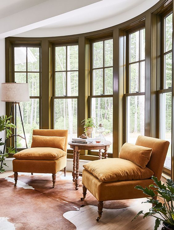a bay window with dark frames, a couple of comfy yellow chairs with pillows, a vintage table and greenery is amazing