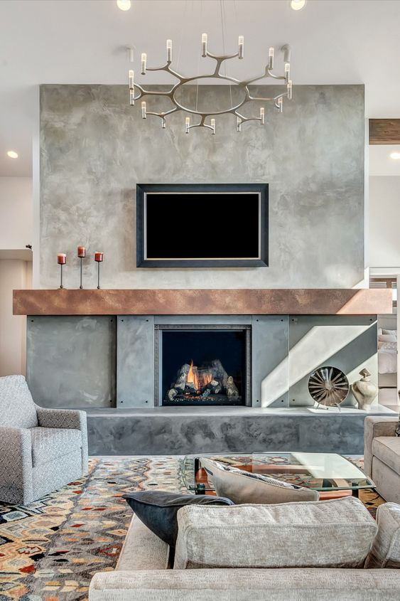 a beautiful and welcoming living room with a concrete fireplace, a wooden mantel, a TV, grey seating furniture, a chic chandelier and a bold printed rug