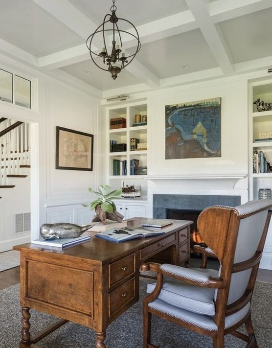 a beautiful vintage home office with a built-in fireplace, built-in shelves, artworks, a vintage heavy desk and a wingback chair is chic