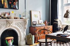 a boho eclectic home office with blue walls, an ornated fireplace, printed layered rugs, a dark desk and a rattan chair, a stained sideboard